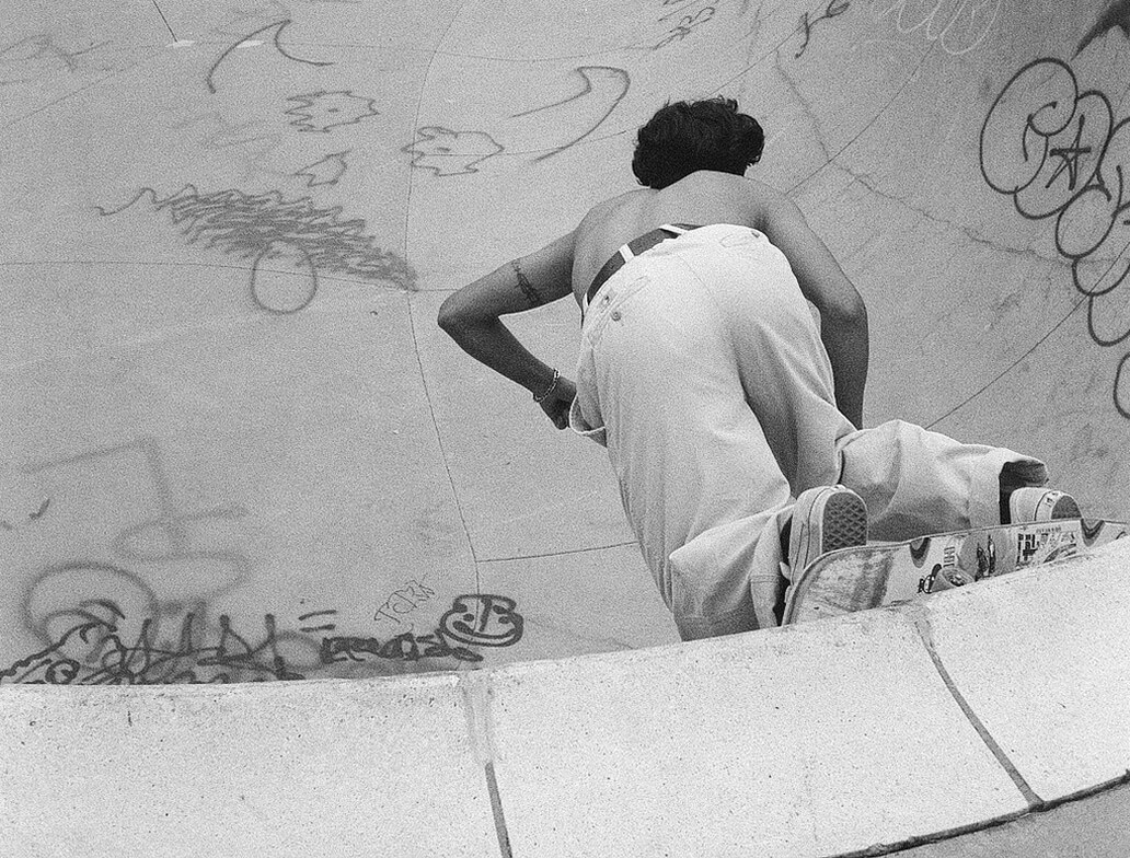 A person in loose pants skateboards in a graffiti-covered concrete skate bowl, captured from behind in black and white.