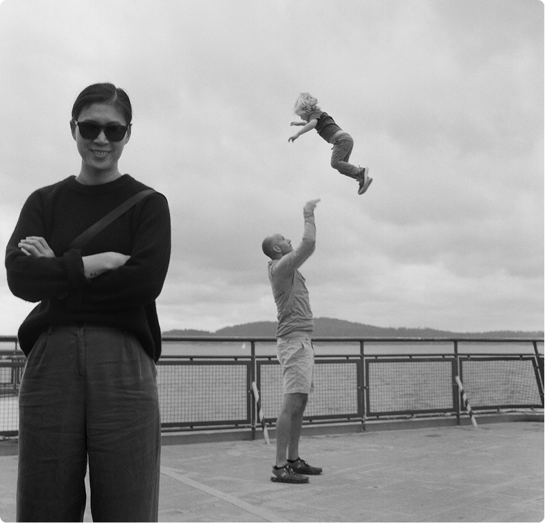 A woman in sunglasses smiles at the camera with arms crossed, while in the background, a man tosses a child playfully into the air on a waterfront pier under a cloudy sky.