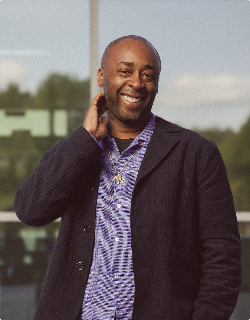 A man wearing a dark jacket and a purple shirt smiles while standing outdoors, with one hand raised to the back of his neck. The background features trees and a modern glass building.