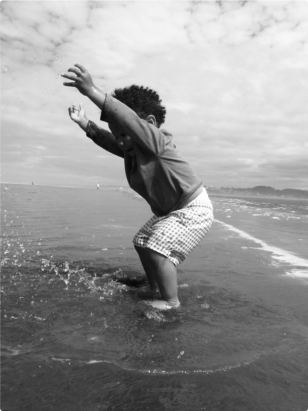 A young child plays joyfully in shallow ocean water, splashing with arms raised. The beach stretches behind under a partly cloudy sky. The image is in black and white.