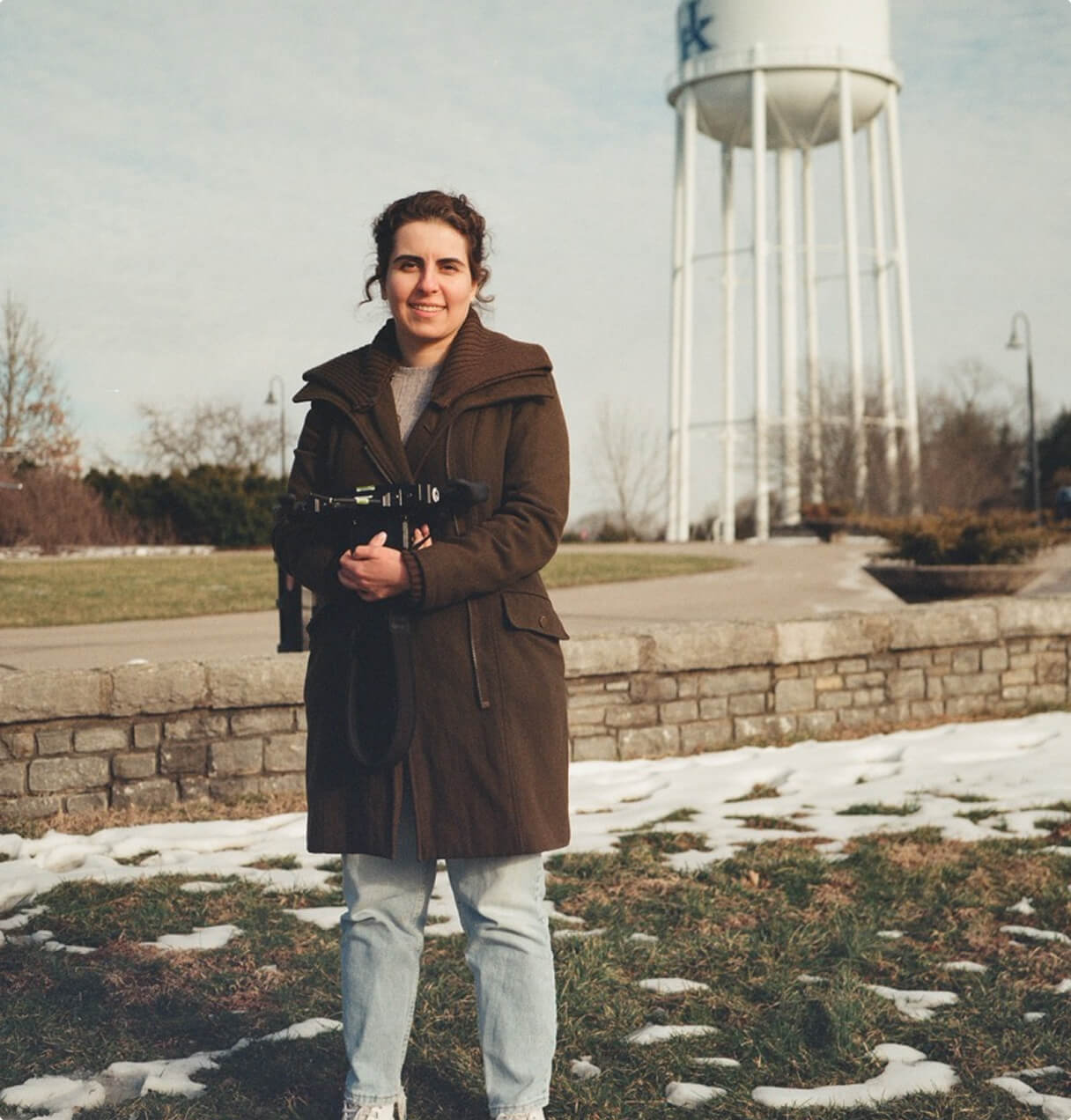 A woman in a brown coat and jeans stands on grass with patches of snow, holding a camera. A stone wall and a white water tower are in the background under a partly cloudy sky.