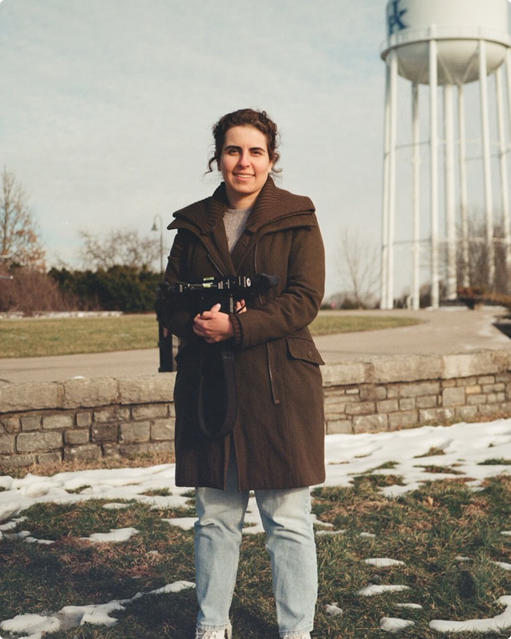 A woman in a brown coat and jeans stands outdoors on a patchy snowy field, holding a camera. There’s a stone path and a tall white water tower in the background under a partly cloudy sky.