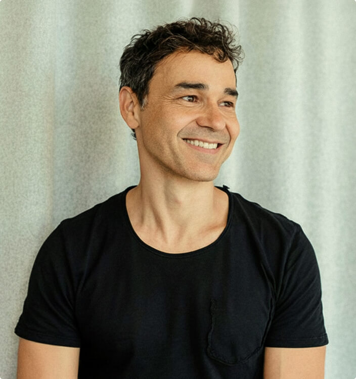 A man with short, curly dark hair wearing a black t-shirt smiles and looks to the side. He is standing in front of a light-colored, textured background.