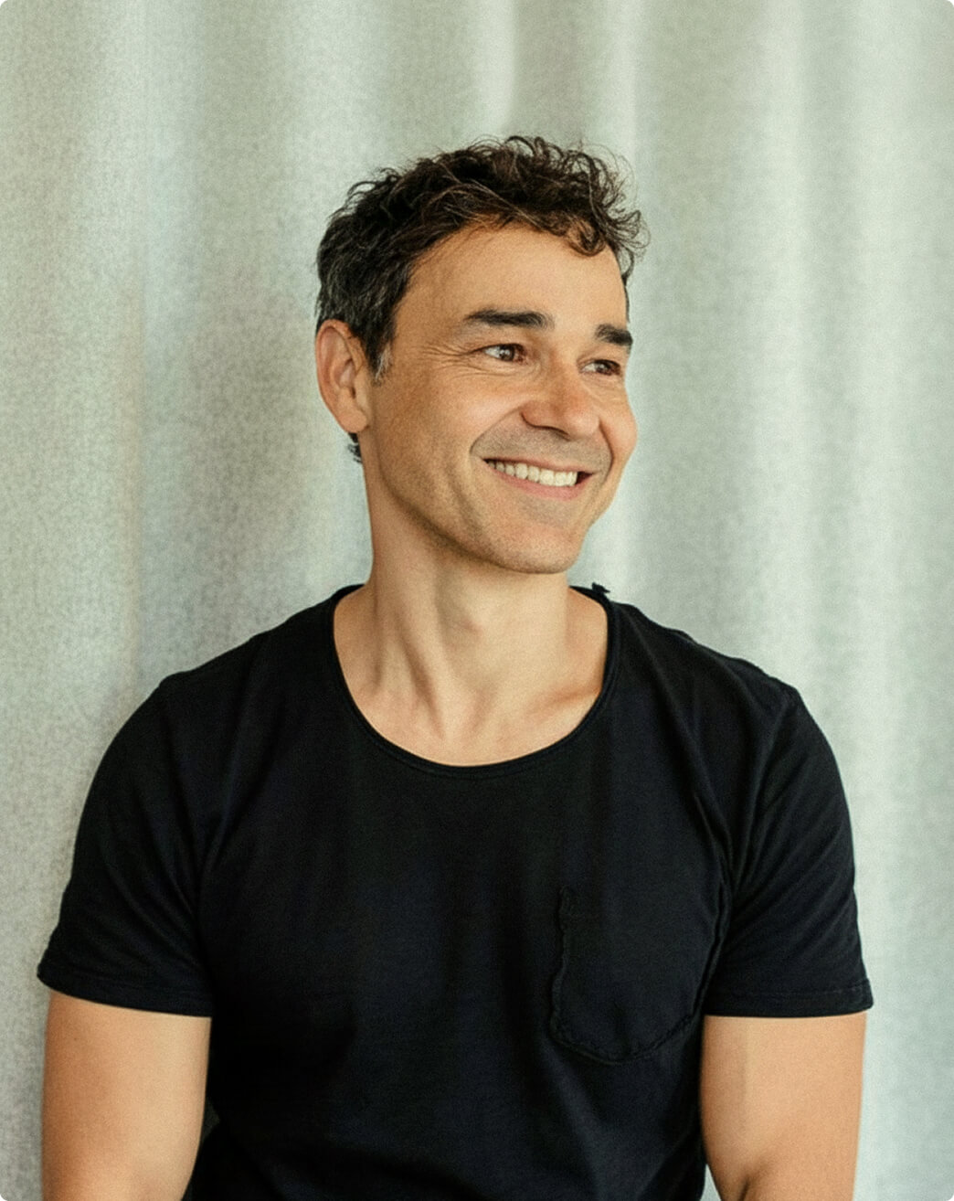 A man with short, curly dark hair and a black T-shirt smiles while looking to the side, standing in front of a light-colored curtain.