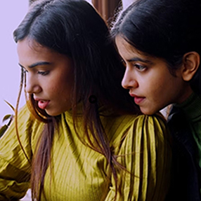 Two people intently looking at a screen, indoors. One wears a yellow top and the other wears a dark top. They appear focused and are positioned closely together, suggesting collaboration or shared interest.