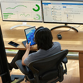 Person sitting at a desk with two large monitors displaying graphs and data analysis tools. They are wearing headphones and holding a tablet. A smartphone and mouse are also on the desk. The setting appears to be a modern office.