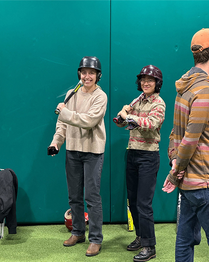 Two people in batting helmets hold baseball bats and smile indoors on artificial turf in front of a teal wall. A person in the foreground, wearing a cap, has their back turned. Baseball equipment is seen on the floor.