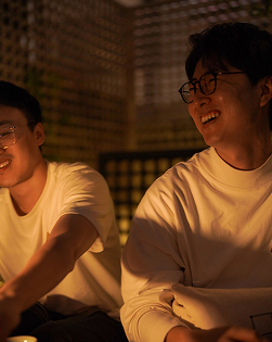 Two people sitting side by side, lit by warm candlelight. They are both wearing glasses and casual white shirts, smiling and looking towards something off-camera. The background has a grid-like pattern.