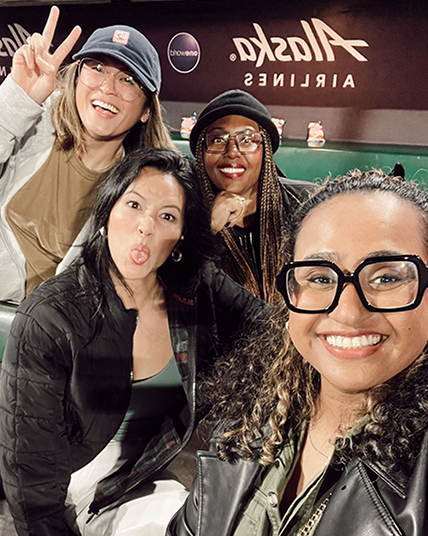 Four people are posing for a selfie, smiling and making playful faces. They are indoors, with an "Alaska Airlines" sign visible in the background.