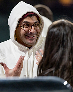 A person in a white hoodie and glasses smiles and gestures with both hands while talking to someone with long dark hair. The scene appears to be candid, capturing an animated moment of conversation.