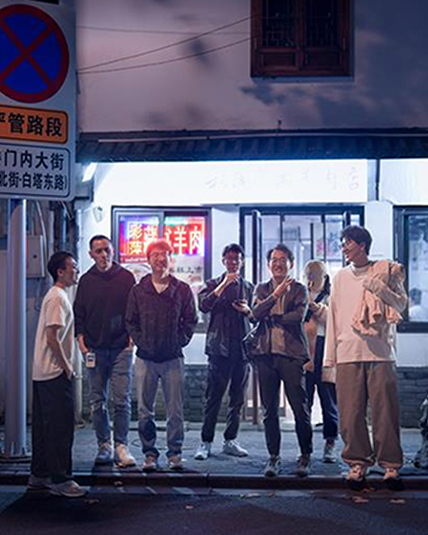 A group of people stand casually on a dimly lit street at night, in front of a building with a neon sign. They appear to be chatting and holding drinks. A street sign is visible on the left.