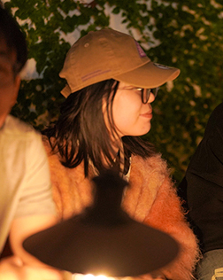 A person wearing a brown cap and glasses sits in a dimly lit outdoor setting, illuminated by a nearby light. The background has green foliage, and they are dressed in a soft, brown sweater.