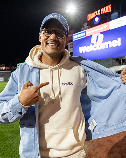 A person wearing glasses and a cap points to their hoodie that reads "copilot" while smiling at a baseball stadium. The scoreboard in the background displays "Welcome!" at Oracle Park.