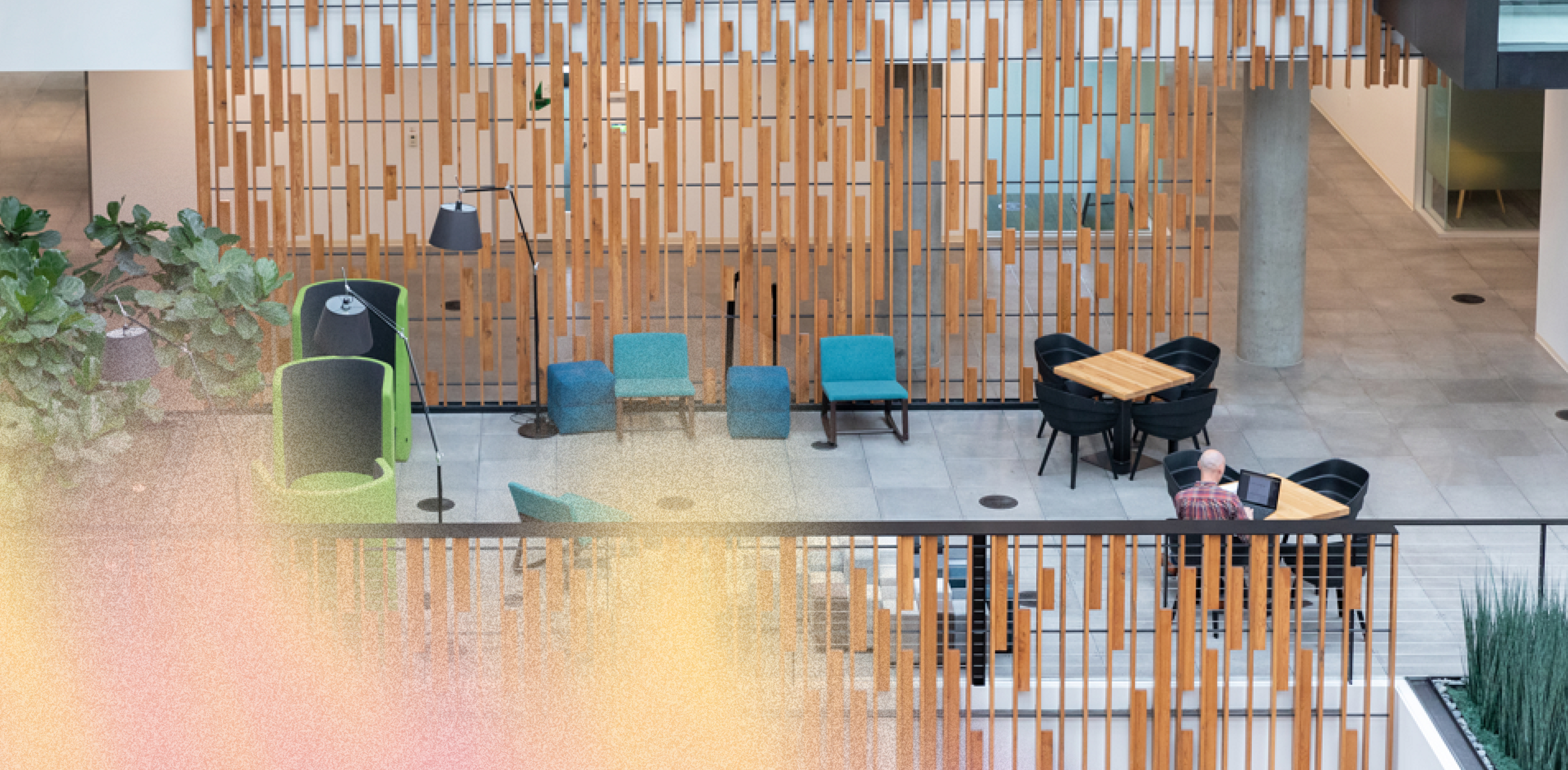 Modern indoor atrium with wooden slat divider, empty blue and green chairs, black tables and chairs, potted plants, and a single person sitting at a table, viewed from above with soft colored light in the foreground.