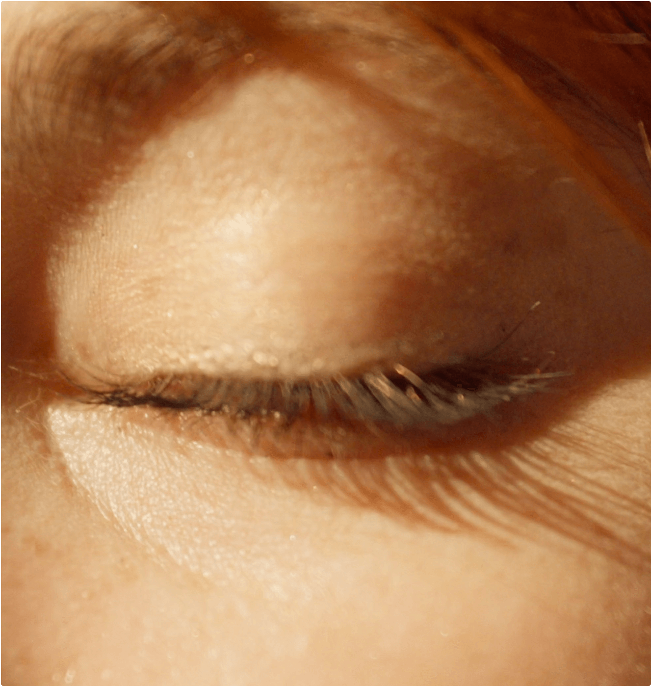 A close-up of a person's closed eye, showing long eyelashes and soft sunlight casting shadows on their skin. The image highlights the texture and warmth of the skin and the natural eyelashes.