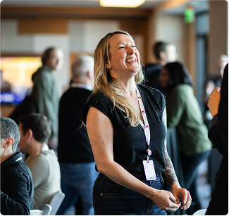 A smiling woman with long blonde hair stands indoors at an event, wearing a black shirt and a lanyard. People are gathered and talking in the background.