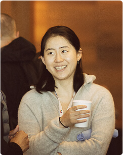 A woman with dark hair smiles while holding a white paper cup. She is wearing a light gray sweater and standing indoors, engaging in conversation with others. The background is softly blurred.