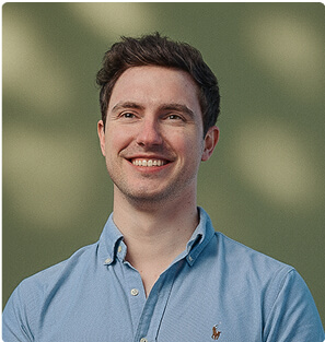 A young man with short brown hair, wearing a light blue button-up shirt, smiles while standing in front of a blurred green background with soft light spots.