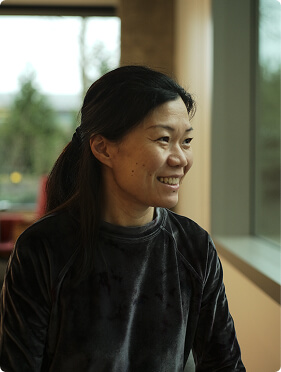 A woman with long black hair tied back is smiling while sitting indoors by a window, wearing a dark, long-sleeved velvet top. Natural light is coming through the window beside her.