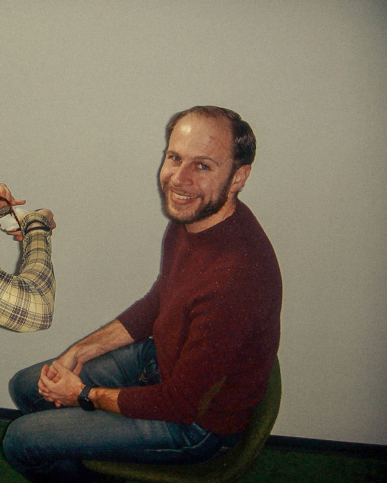 A man with a beard, wearing a maroon sweater and jeans, sits sideways on a green chair and smiles at the camera. Another person’s hand holding glasses is visible on the left side of the image.
