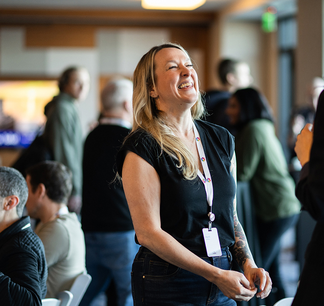 A woman with long blonde hair, wearing a black t-shirt and name badge, stands smiling and laughing in a busy room filled with people engaged in conversation.