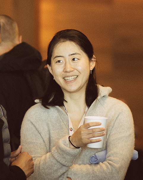 A woman with dark hair, wearing a light gray sweater, smiles while holding a white paper cup. She stands in a warmly lit indoor setting with other people blurred in the background.