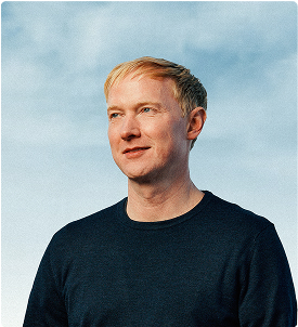 A man with short blond hair in a dark sweater stands outdoors against a blue sky, looking slightly to the side with a neutral expression.
