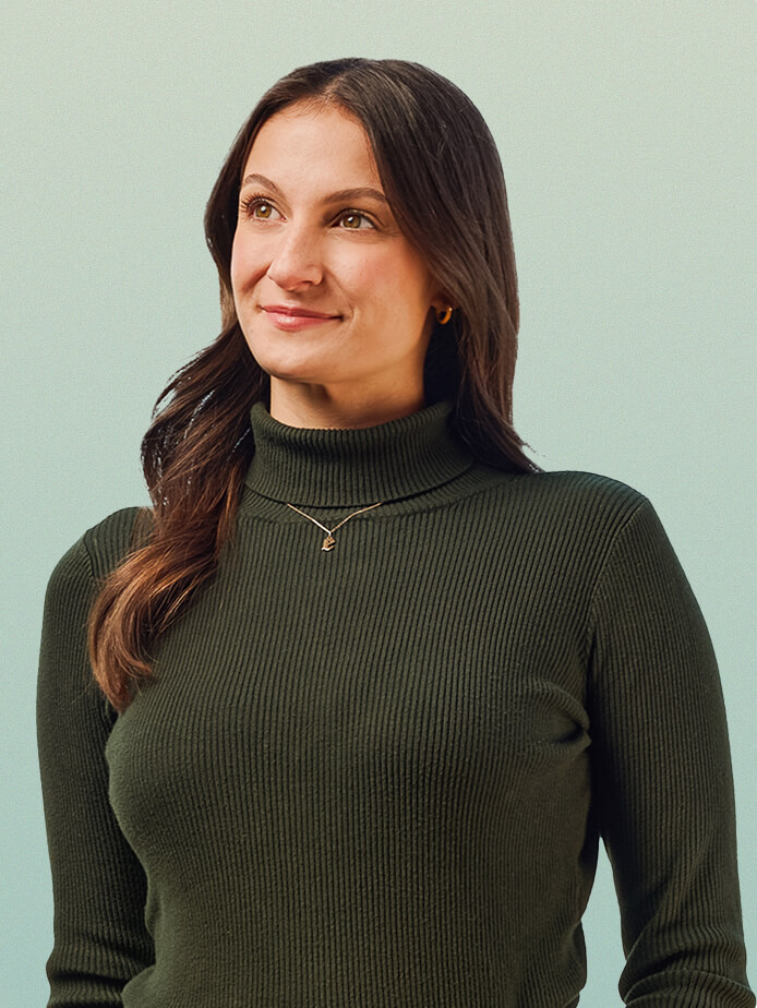 A woman with long brown hair wearing a dark ribbed turtleneck sweater and a gold necklace, standing against a light blue background and looking slightly to her left with a gentle smile.