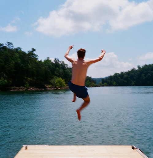 A person in blue swim trunks jumps off a wooden dock into a lake, arms raised and knees bent, with trees and a blue sky with clouds in the background.