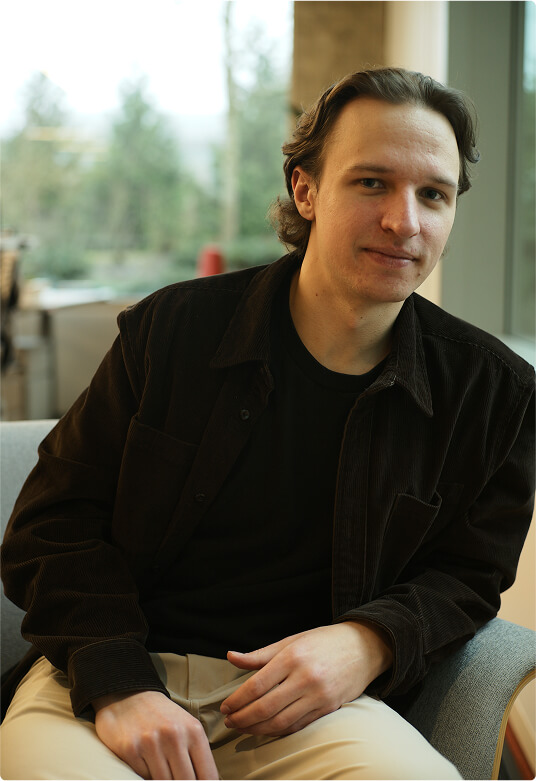 A man with light skin and brown hair, wearing a black shirt and brown jacket, sits on a light-colored chair indoors, looking at the camera with a slight smile. A window with a blurred outdoor view is behind him.