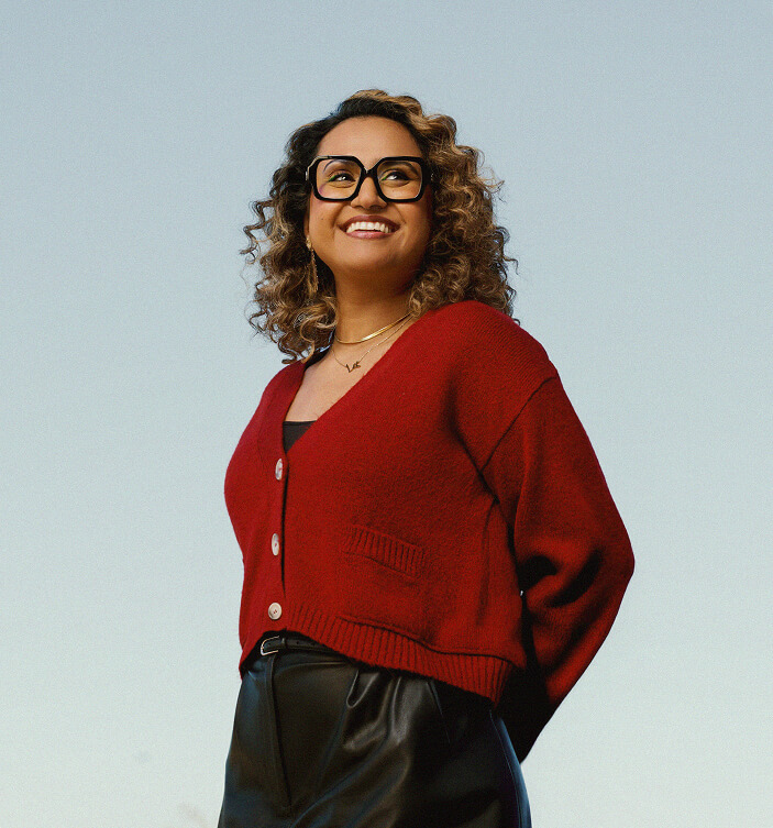 A woman with curly hair and glasses smiles, looking upward. She wears a red cardigan over a black top and black pants, standing outdoors with a clear blue sky in the background.