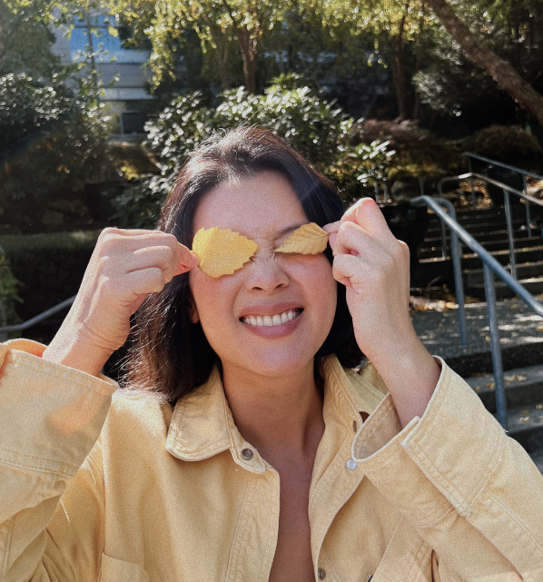 A woman in a light yellow jacket smiles while holding two yellow leaves over her eyes, standing outdoors with trees and stairs in the background.
