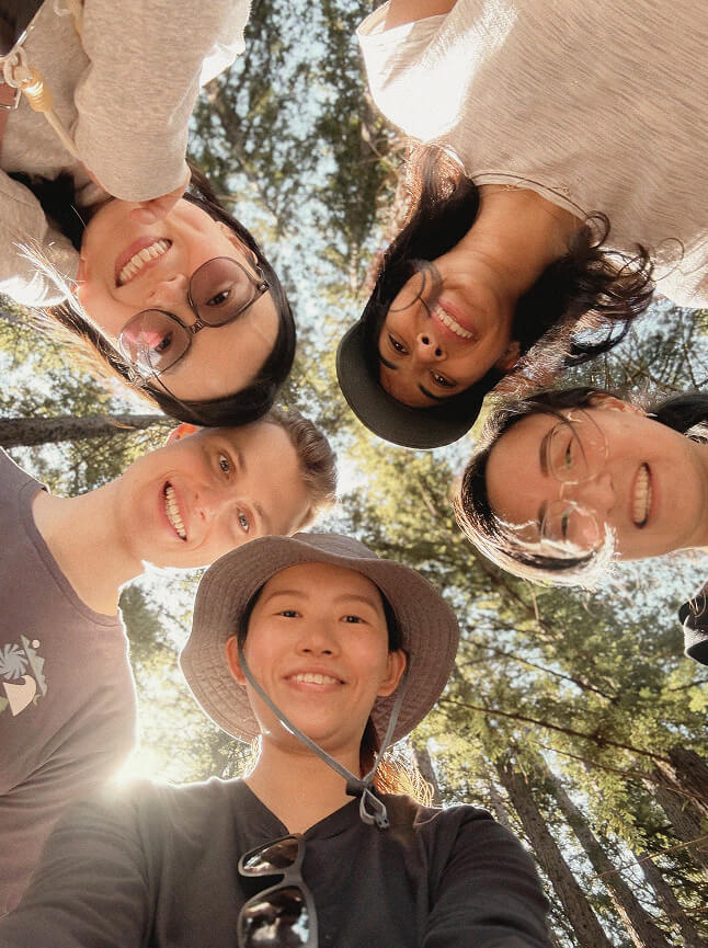 Five people stand in a circle outdoors, smiling down at the camera surrounded by tall trees and sunlight. They wear casual clothes, glasses, and hats, creating a cheerful, friendly atmosphere.