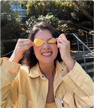 A woman in a yellow jacket smiles while holding two yellow leaves over her eyes outdoors, with stairs and greenery in the background.