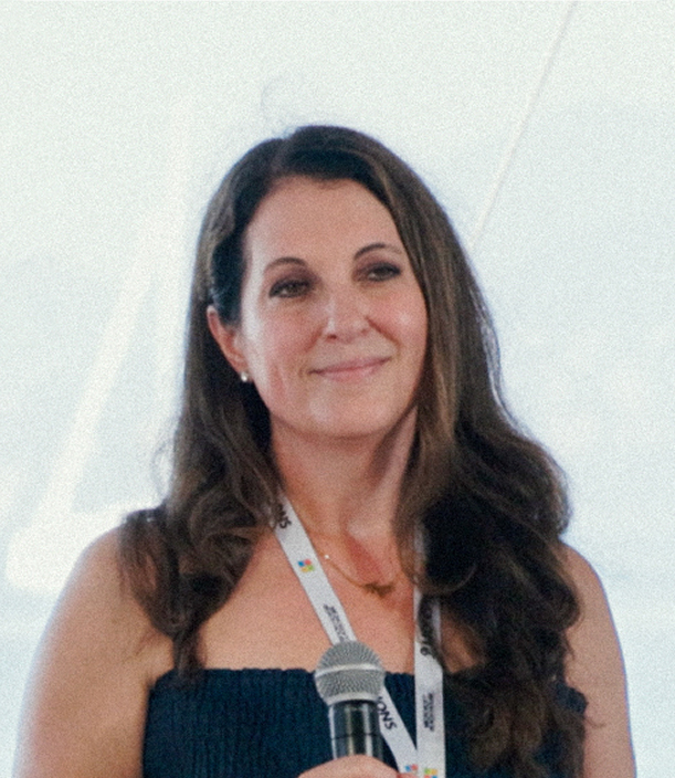 A woman with long brown hair, wearing a dark strapless top and a lanyard, smiles while holding a microphone against a light background.