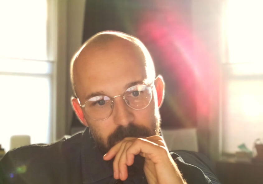 A person with glasses and a beard rests their chin on their hand, looking thoughtful. Sunlight streams in from large windows behind them, creating a bright, soft glow and lens flare in the image.