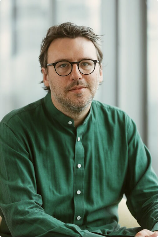 A man with glasses and dark hair, wearing a green button-up shirt, sits indoors in front of large windows with a neutral expression.