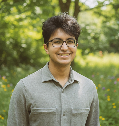 A young person with short dark hair and glasses smiles while standing outdoors in a sunlit, green area with trees and wildflowers in the background. They are wearing a light gray collared shirt.