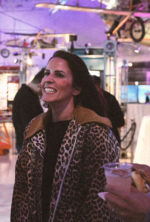 A smiling woman wearing a leopard print coat stands indoors at a lively event, with colorful lights and people in the background. A hand holding a drink is visible in the foreground.