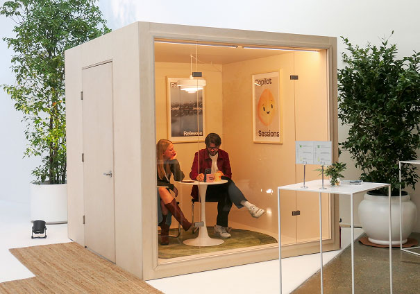 Two people sit at a small round table inside a modern, glass-walled office pod. They appear to be talking and writing. The pod is surrounded by potted plants, and posters are visible on its walls.