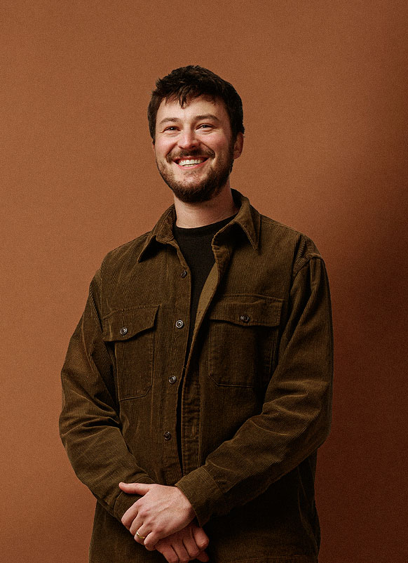 A man with short dark hair and a beard smiles while standing against a plain brown background. He is wearing a brown corduroy jacket over a black shirt, with his hands folded in front of him.