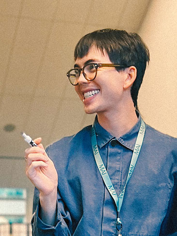 A person wearing glasses and a blue shirt with a lanyard smiles while holding a marker. The background appears to be indoors, possibly in an office or classroom setting.