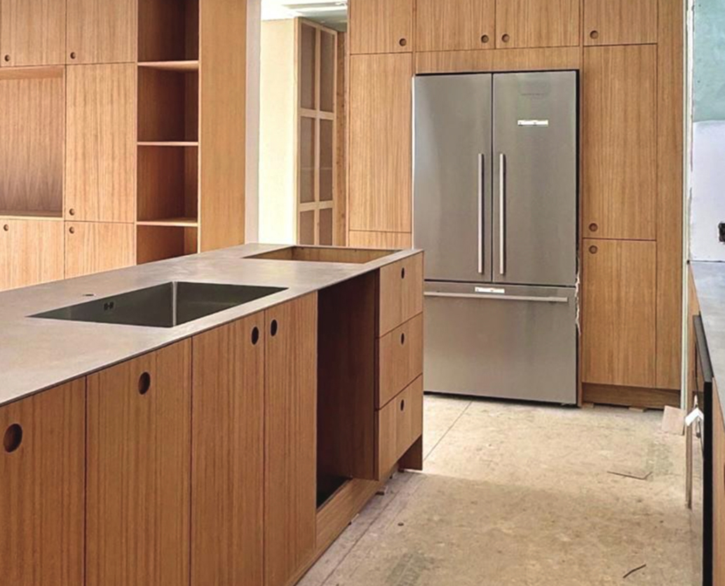 Modern kitchen with light wood cabinets, a large island featuring a built-in sink, and a stainless steel French door refrigerator. The kitchen appears unfinished with no flooring or appliances installed yet.