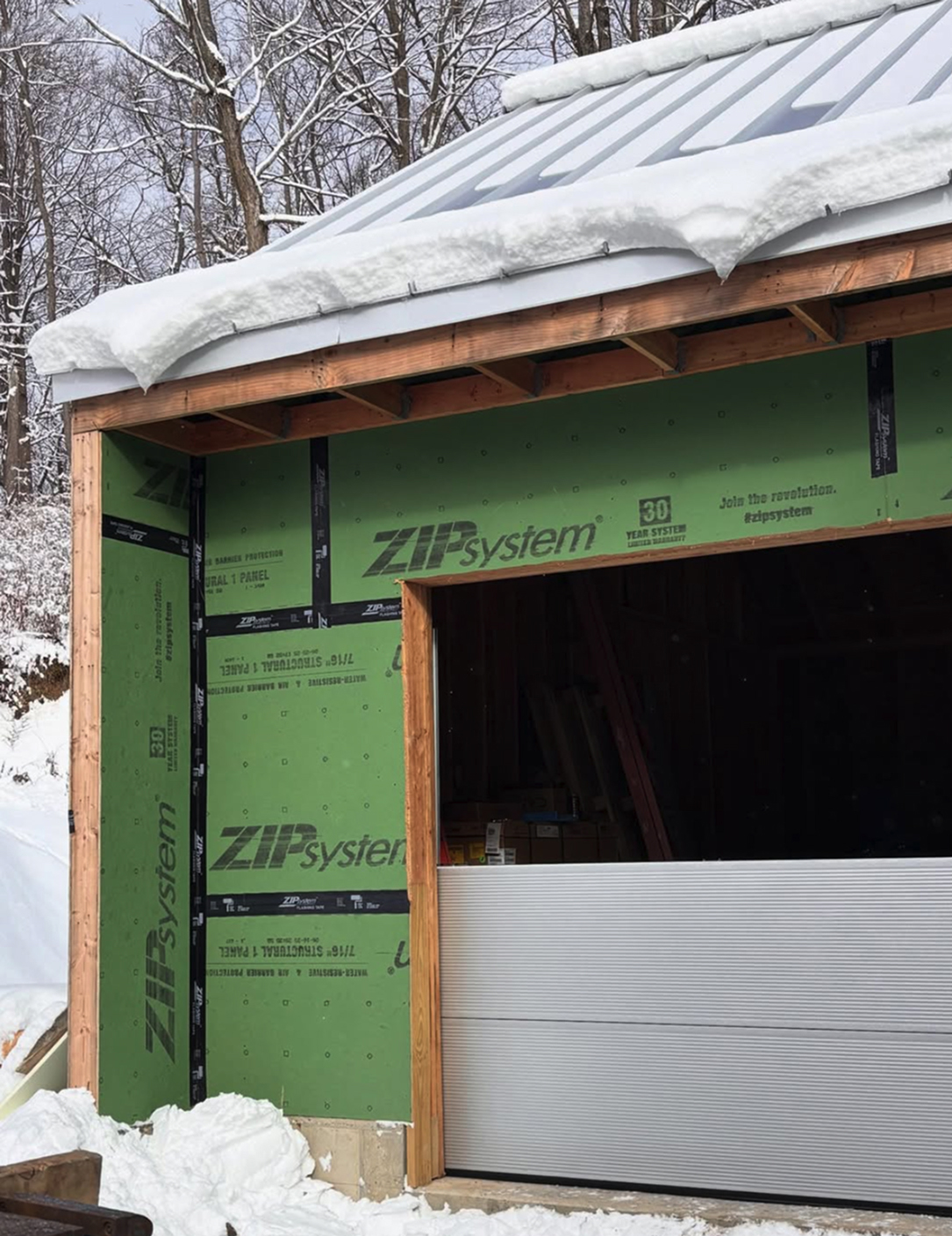 A partially constructed building with green ZIP System sheathing and an open garage door under a snow-covered roof. Snow is piled on the roof and ground, and trees are visible in the background.