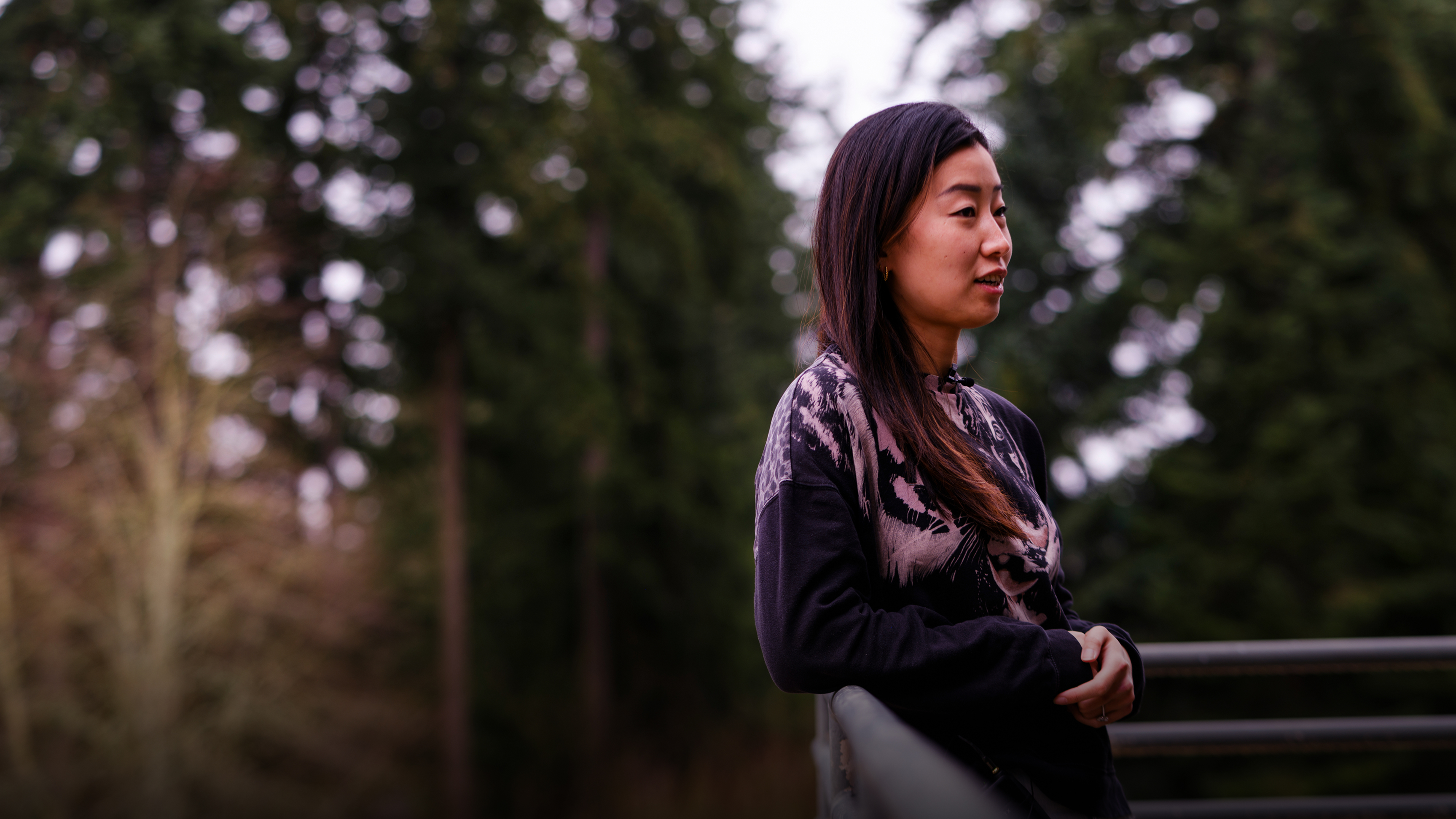 A woman with long dark hair stands outdoors by a metal railing, facing right. She wears a black and beige patterned sweater, with tall evergreen trees blurred in the background.