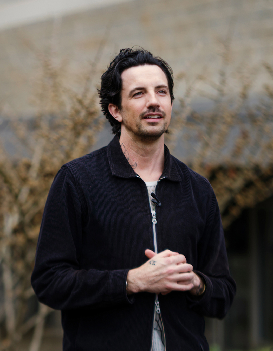 A man with dark wavy hair and facial hair stands outdoors, wearing a black jacket over a striped shirt. He is clasping his hands and looking slightly to the side, with blurred trees and a building in the background.