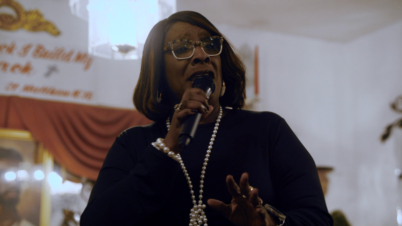 A woman wearing glasses and a pearl necklace sings passionately into a microphone at an indoor event, with a blurred background featuring warm colors and decorations.