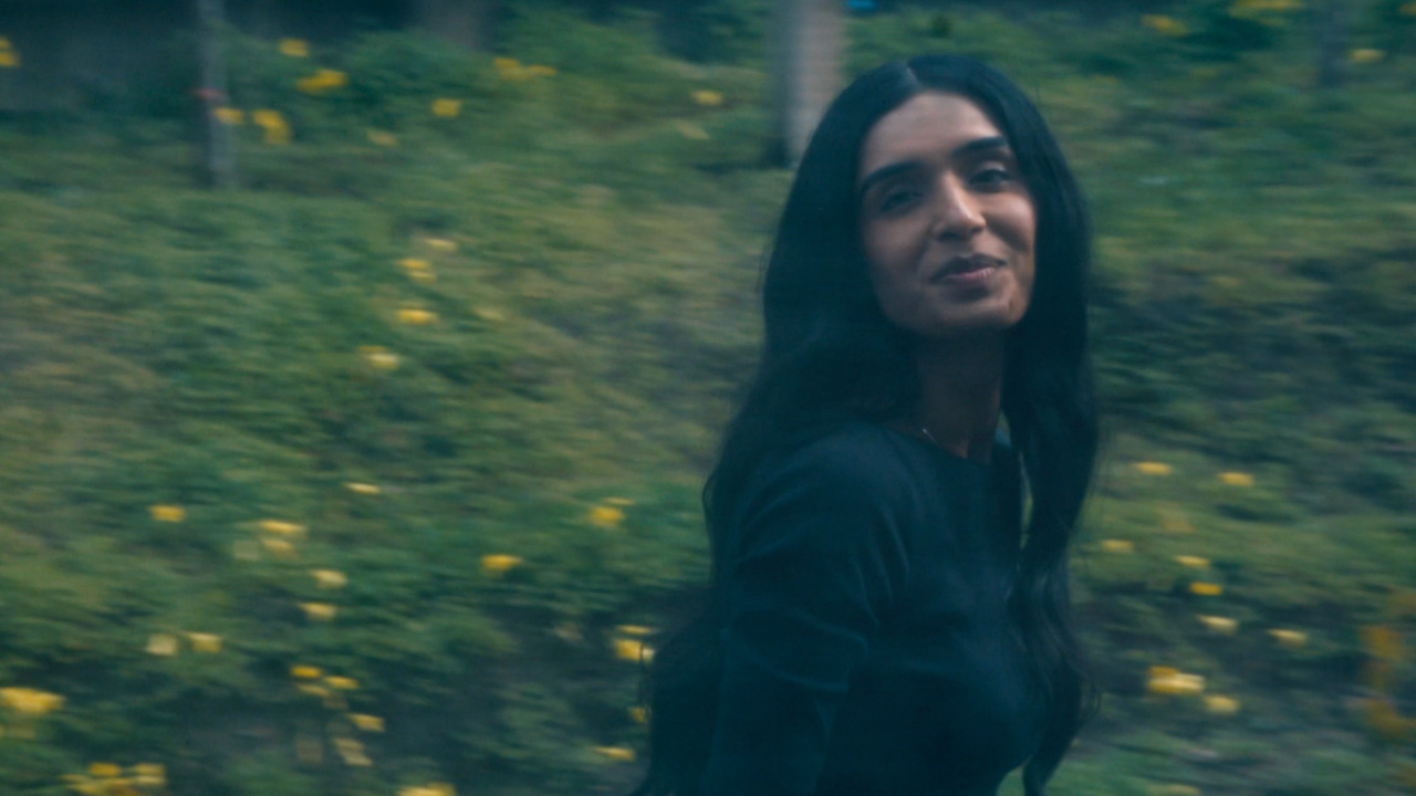 A woman with long dark hair, wearing a dark top, stands outdoors in front of greenery and yellow flowers, looking back over her shoulder with a slight smile.