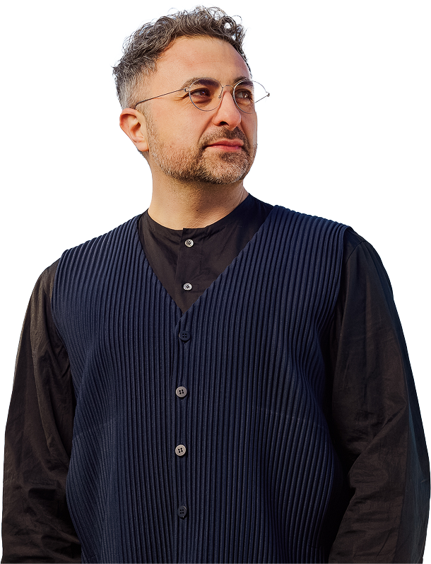 A man with short curly hair, glasses, and a trimmed beard is wearing a pleated navy vest over a black shirt, looking slightly to the side against a plain white background.