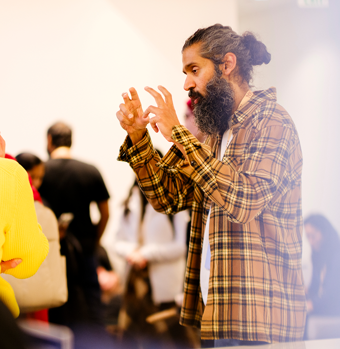 A man with a long beard and tied-back hair, wearing a yellow plaid shirt, gestures animatedly with his hands while speaking to someone in a crowded indoor space.